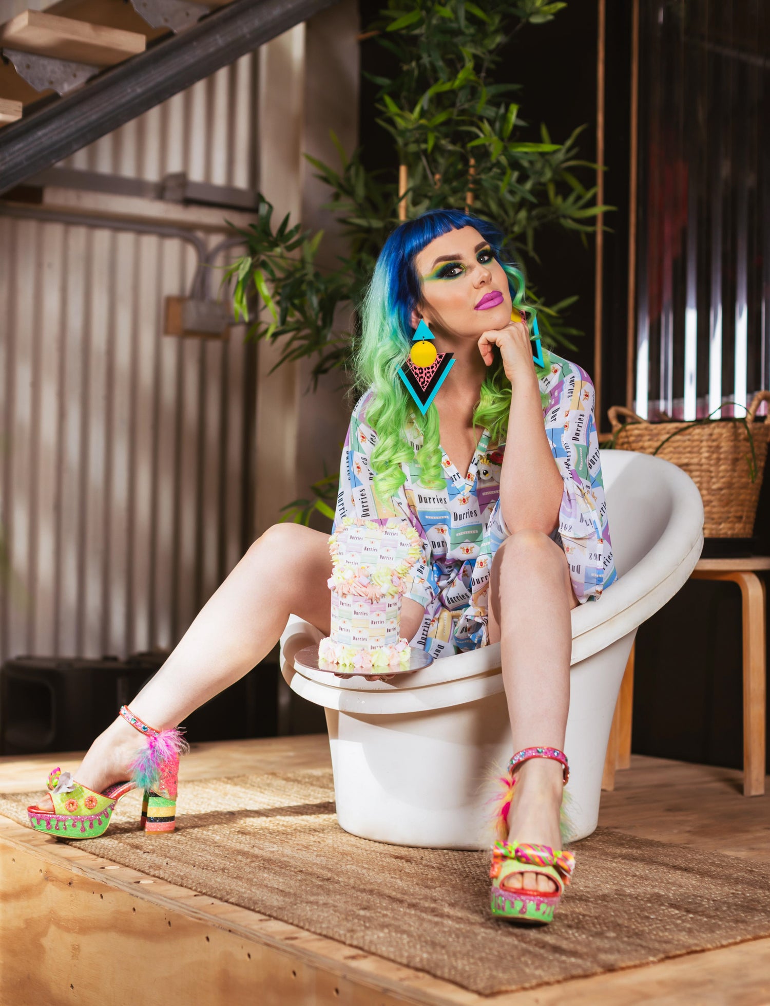 Woman with blue hair sits on vintage white seat holding a cake that matches her dress