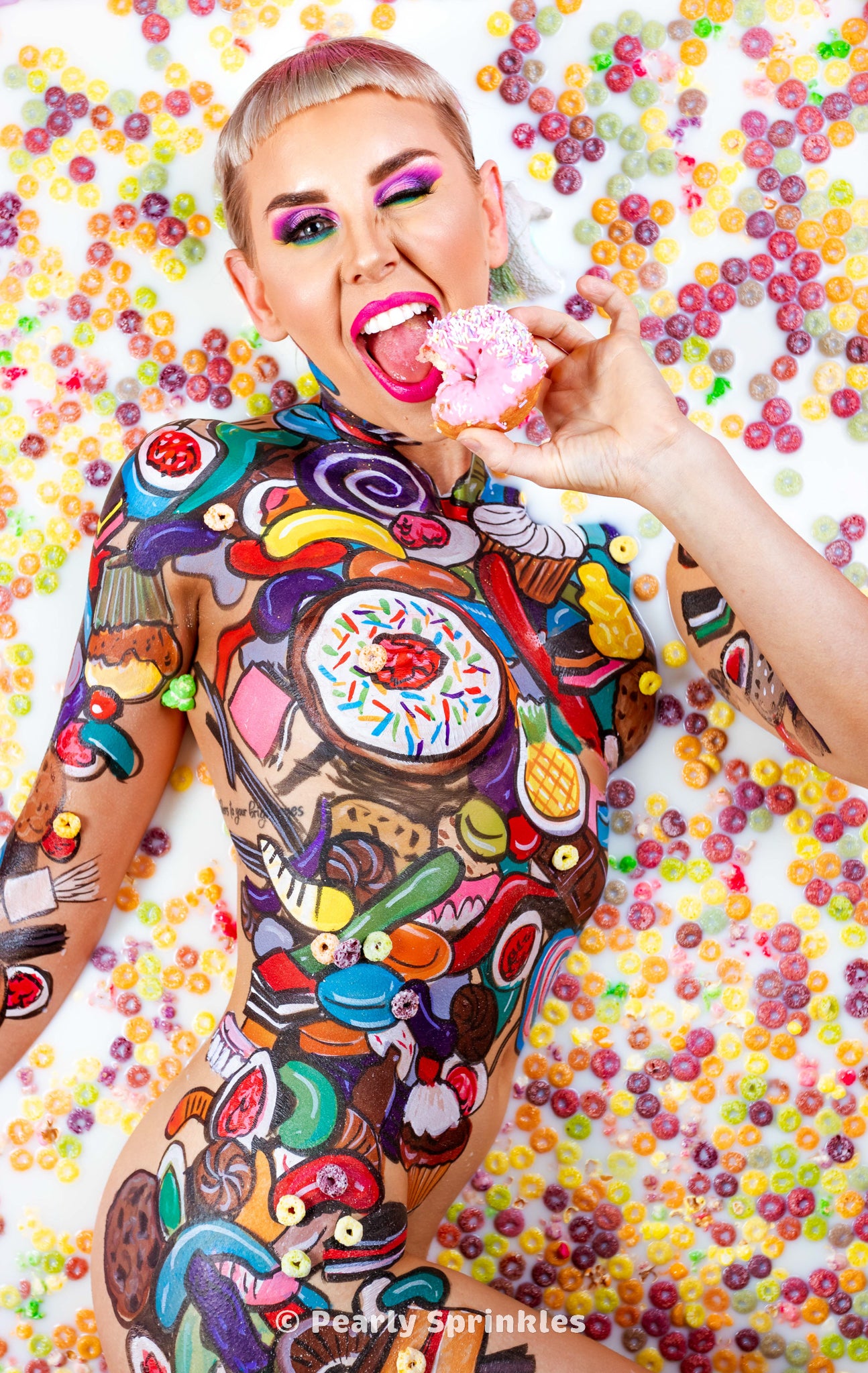 A woman with colorful makeup winks while taking a bite of a donut. She is nude but covered in body painted images sweets and candy, lying in a pool of milk and rainbow cereal.