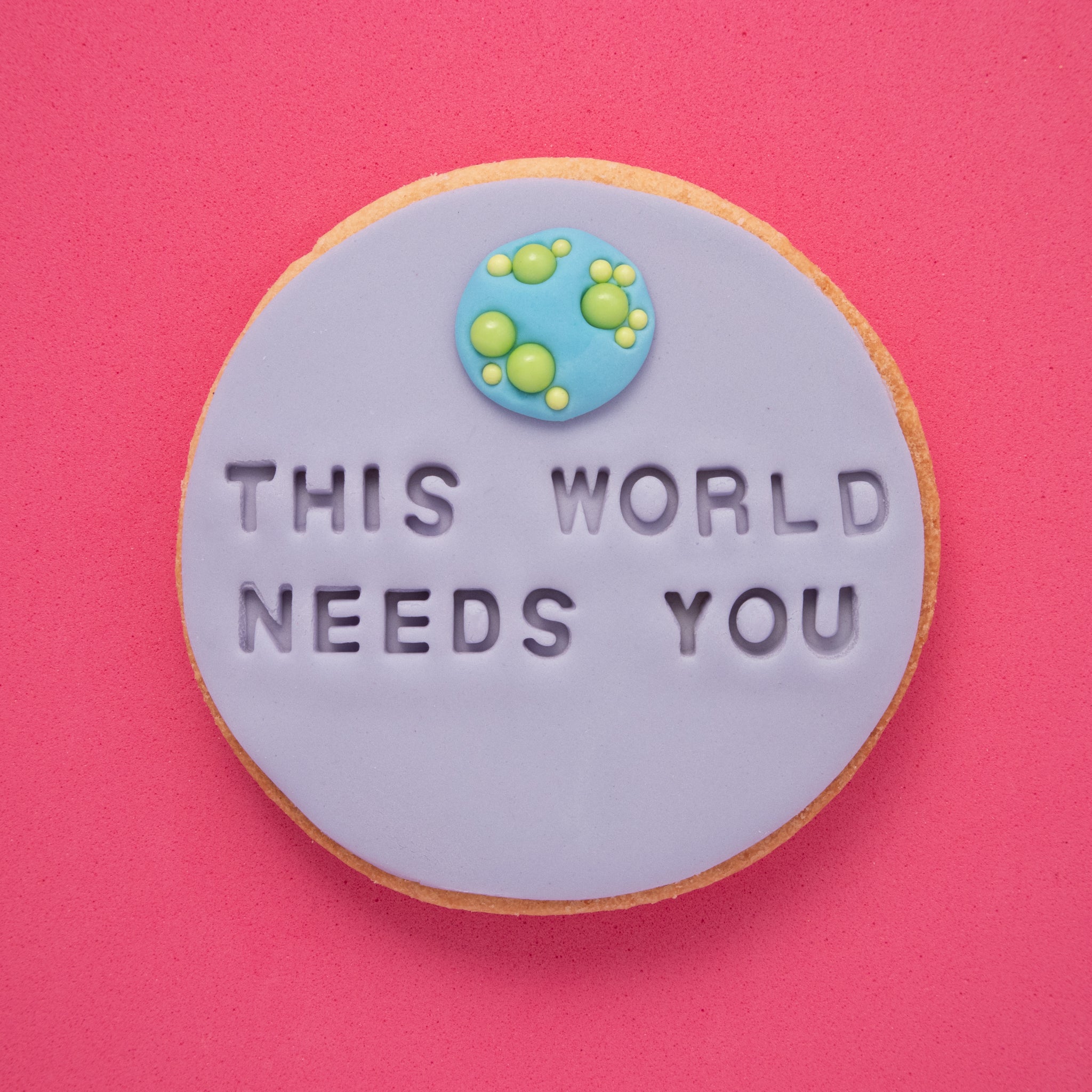 sugar cookie with purple fondant embossed with 'this world needs you' on a pink background