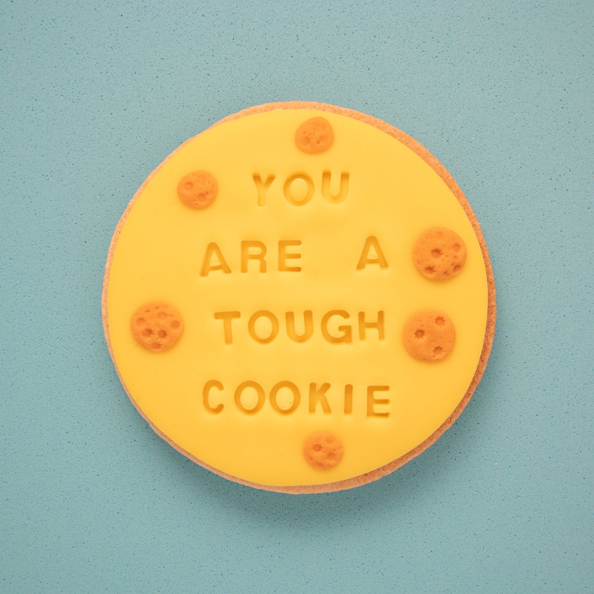 sugar cookie with yellow fondant embossed with 'you are a tough cookie' on a blue background